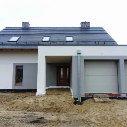 Nowoczesny dom jednorodzinny z jasną elewacją, szarą stolarką okienną i dwoma segmentowymi bramami garażowymi, w trakcie prac wykończeniowych wokół budynku.