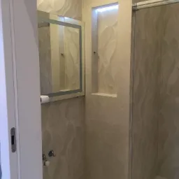 Nowoczesna łazienka z beżowymi płytkami o falistym wzorze, podświetlana wnęka w ścianie i lustro z oświetleniem LED, widoczny fragment kabiny prysznicowej.