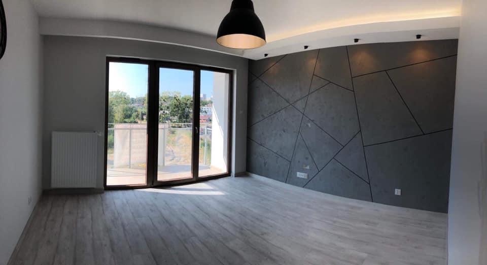 Pusty pokój po remoncie z panelami ściennymi w odcieniach szarości z geometrycznym wzorem, drewnopodobną podłogą i dużym oknem balkonowym.