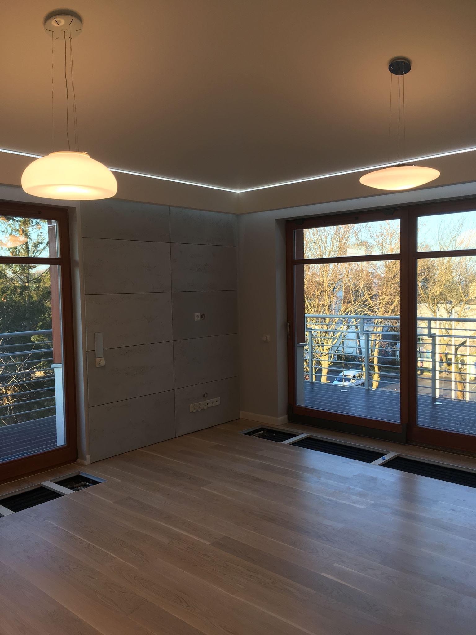 Jasny parkiet ułożony w przestronnym pokoju z dużymi oknami balkonowymi i nowoczesnym oświetleniem sufitowym, widoczne niedokończone otwory wentylacyjne w podłodze.