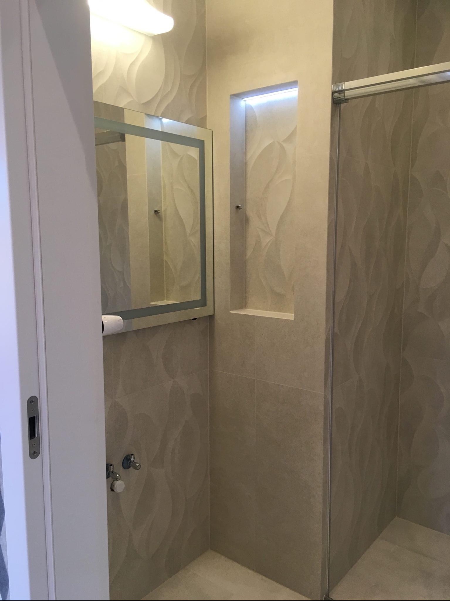 Nowoczesna łazienka z beżowymi płytkami o falistym wzorze, podświetlana wnęka w ścianie i lustro z oświetleniem LED, widoczny fragment kabiny prysznicowej.