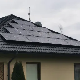 Panele słoneczne zamontowane na dachu domu z czarną dachówką, widoczna antena i satelita na dachu, pochmurne niebo w tle.