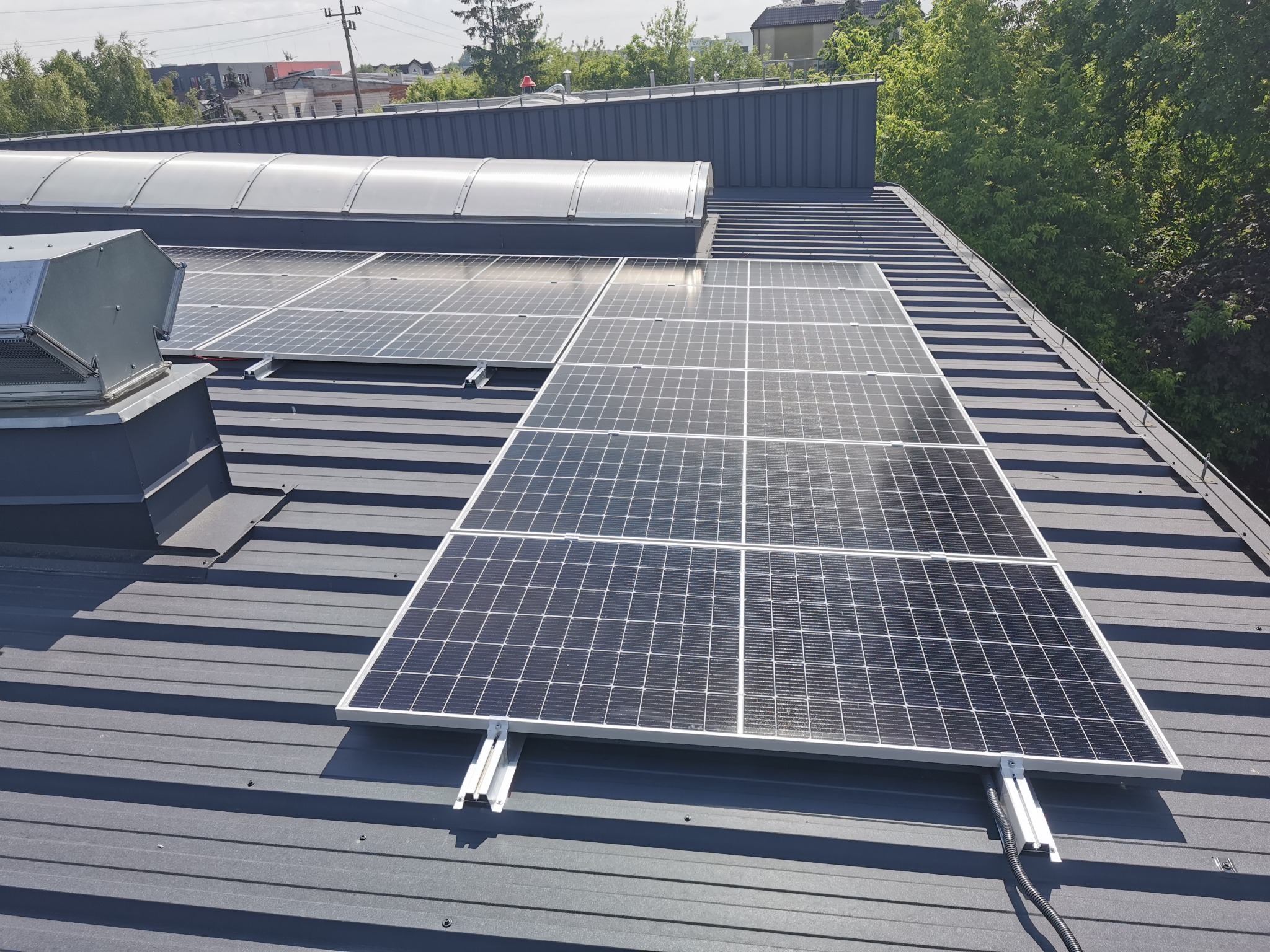 Panele słoneczne zamontowane na metalowym dachu z widocznymi elementami mocującymi i przewodami, oświetlone jasnym światłem słonecznym, z budynkami i zielenią w tle.