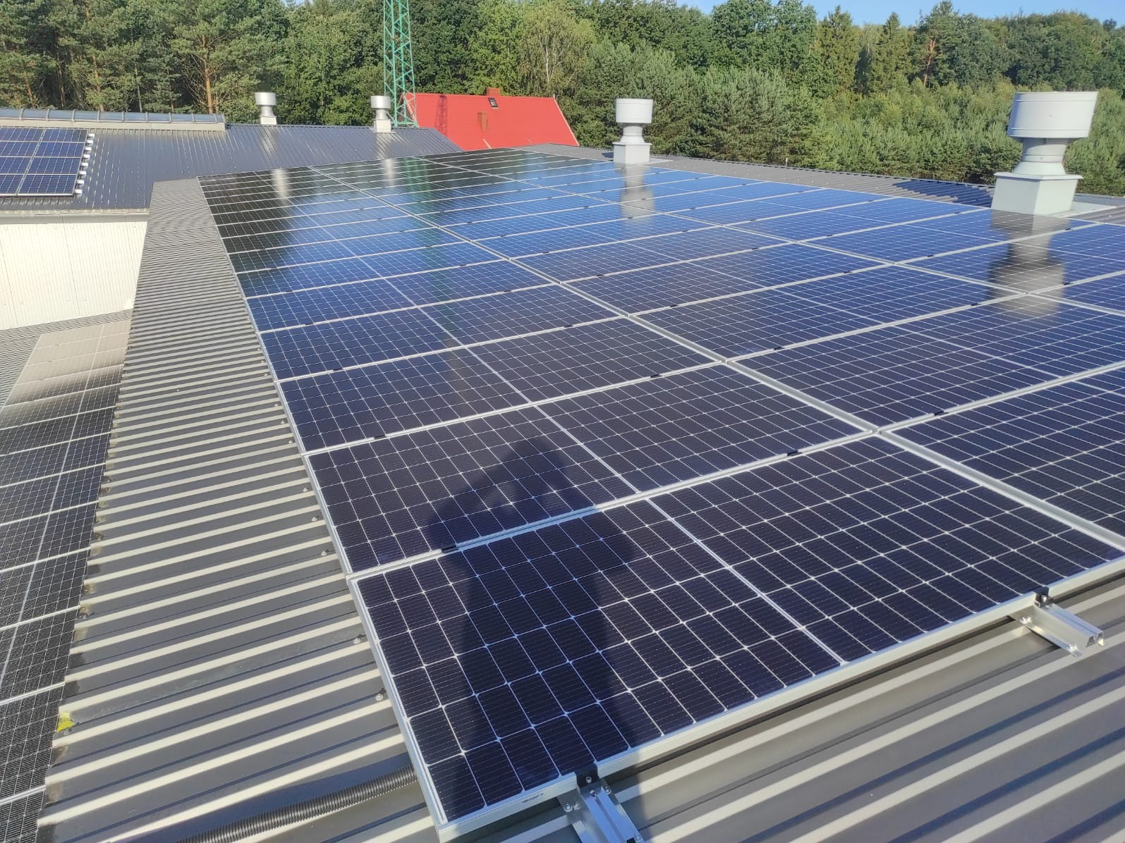 Rząd paneli słonecznych na metalowym dachu przemysłowym z widocznymi elementami montażowymi, wentylacją i zielenią w tle.
