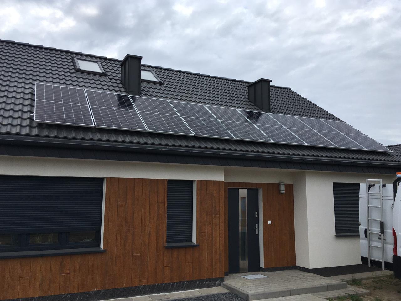 Panele słoneczne na dachu domu jednorodzinnego z ciemną dachówką, widoczne okna dachowe i komin, częściowo zasłonięte rolety w oknach na elewacji.