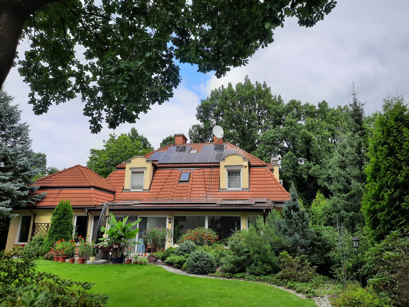 Dom jednorodzinny z czerwoną dachówką i panelami fotowoltaicznymi na dachu, otoczony zielenią i drzewami, widoczna antena satelitarna.