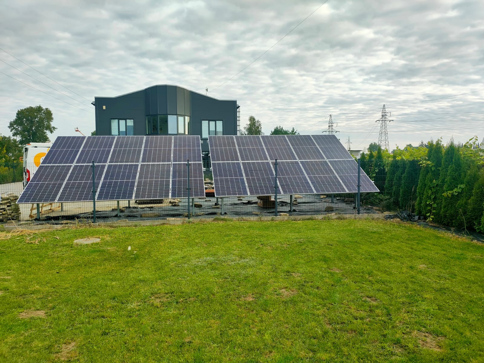 Dwa rzędy paneli słonecznych ustawione na trawniku przed nowoczesnym domem z ciemną elewacją, widoczne elementy konstrukcyjne i tymczasowe zabezpieczenia.