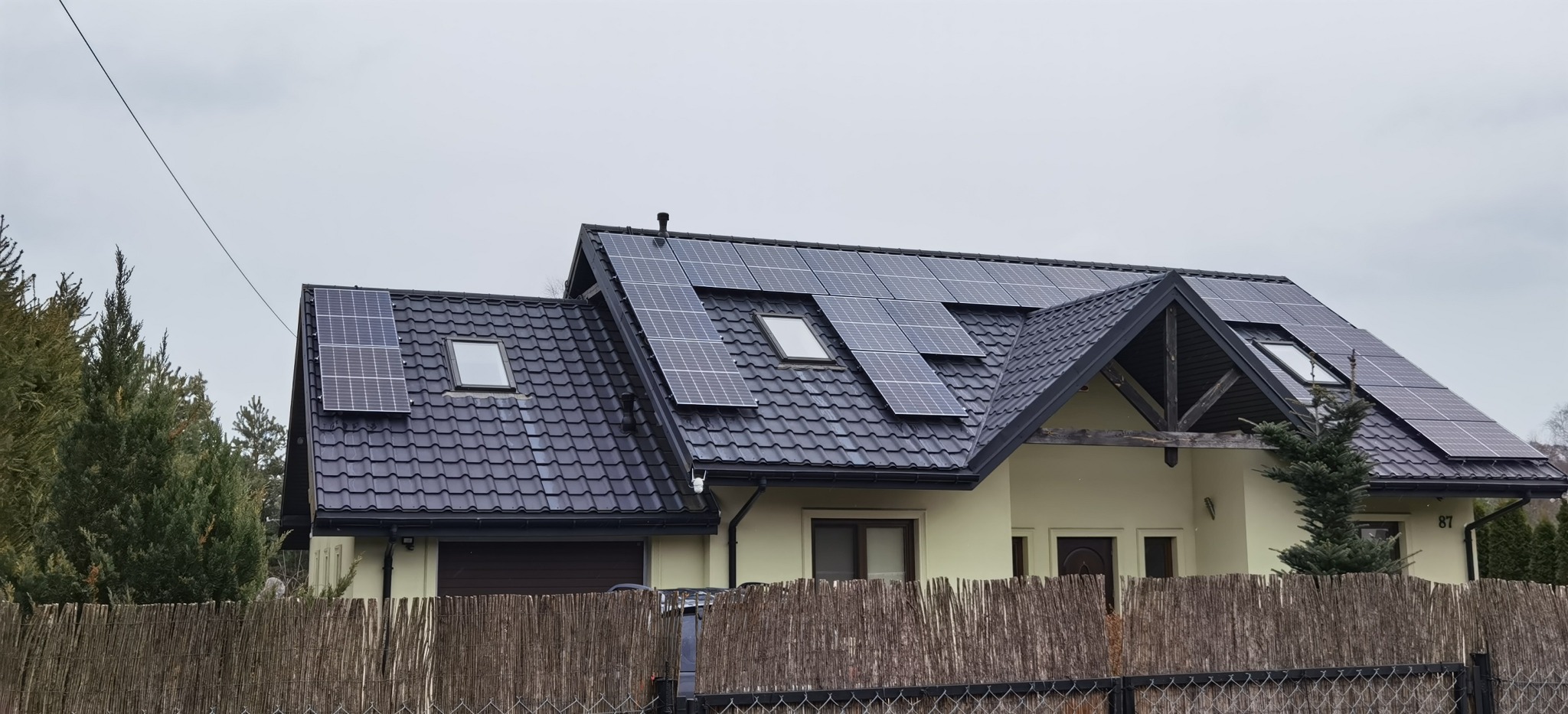 Dom jednorodzinny z ciemną dachówką, na której zamontowano panele słoneczne, widoczne okna dachowe i numer domu 87.