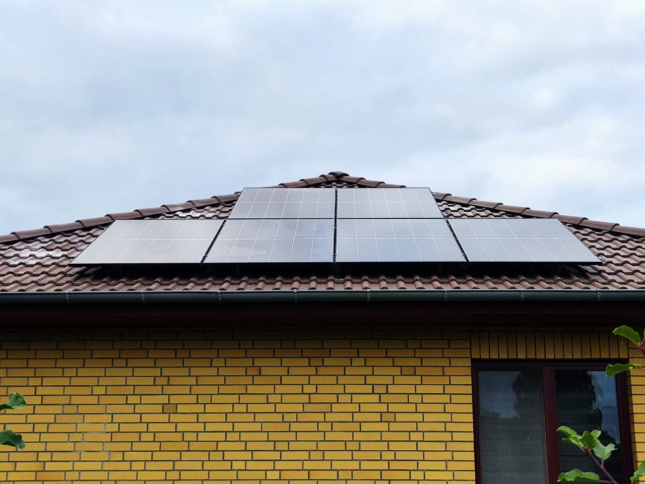 Panele słoneczne na dachu domu z brązową dachówką, widok z dołu na żółtą ceglaną ścianę budynku i fragment okna.