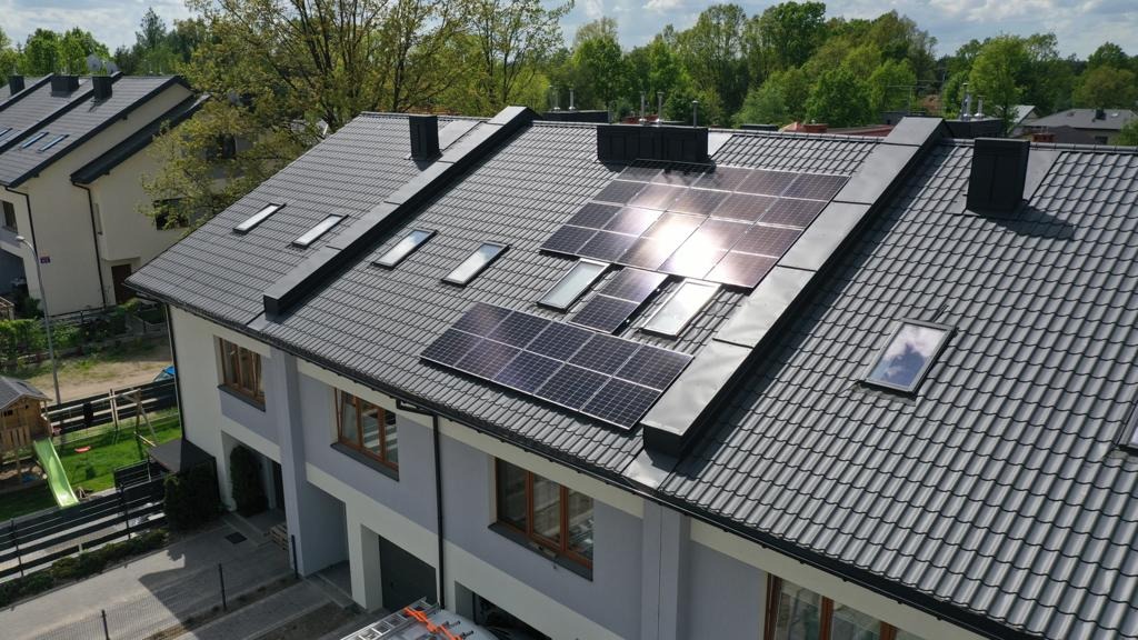 Panele słoneczne na dachu szeregowca z szarej dachówki, częściowo odbijające słońce, z oknami dachowymi, widok z góry na dom i ogród.