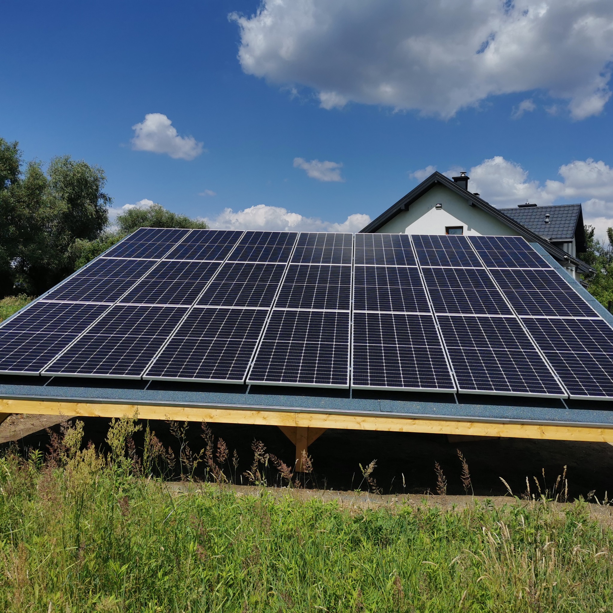 Panel słoneczny na drewnianej konstrukcji z widokiem na dom i błękitne niebo z chmurami.