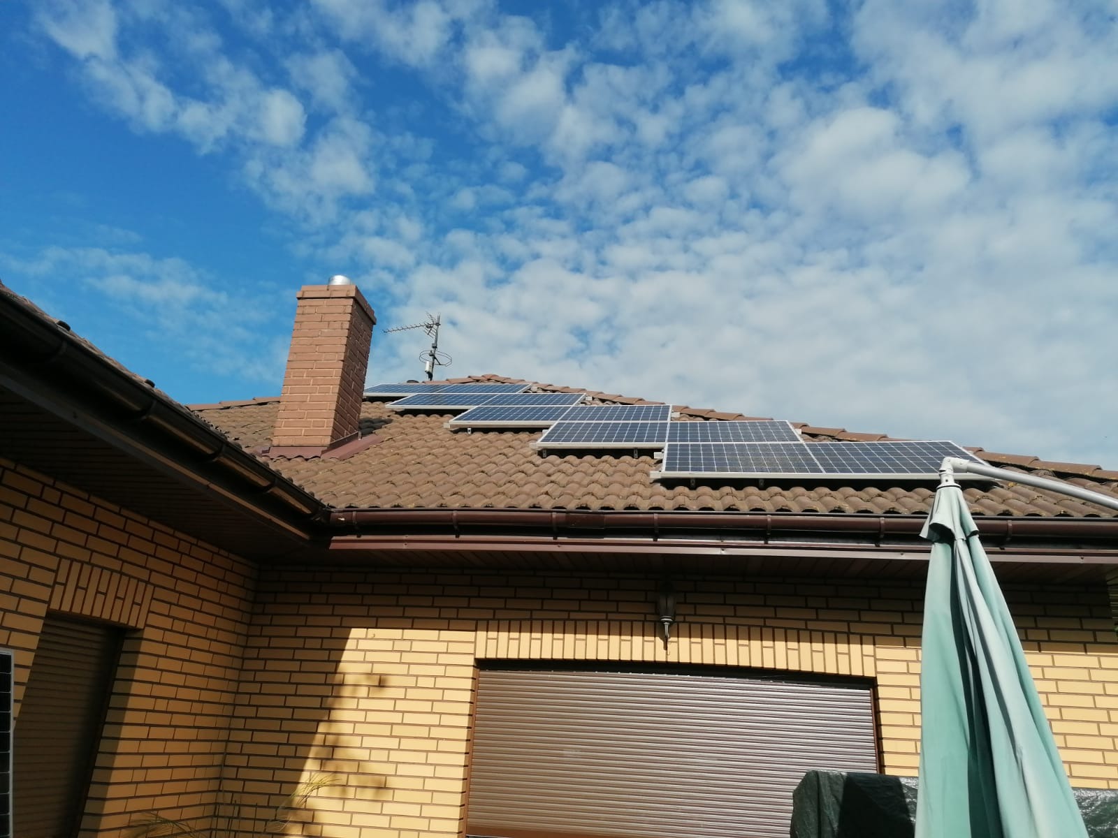Panele słoneczne zamontowane na brązowym, spadzistym dachu domu z cegły, widoczne z dołu pod kątem, z kominem i anteną na dachu oraz fragmentem rozłożonego parasola ogrodowego.