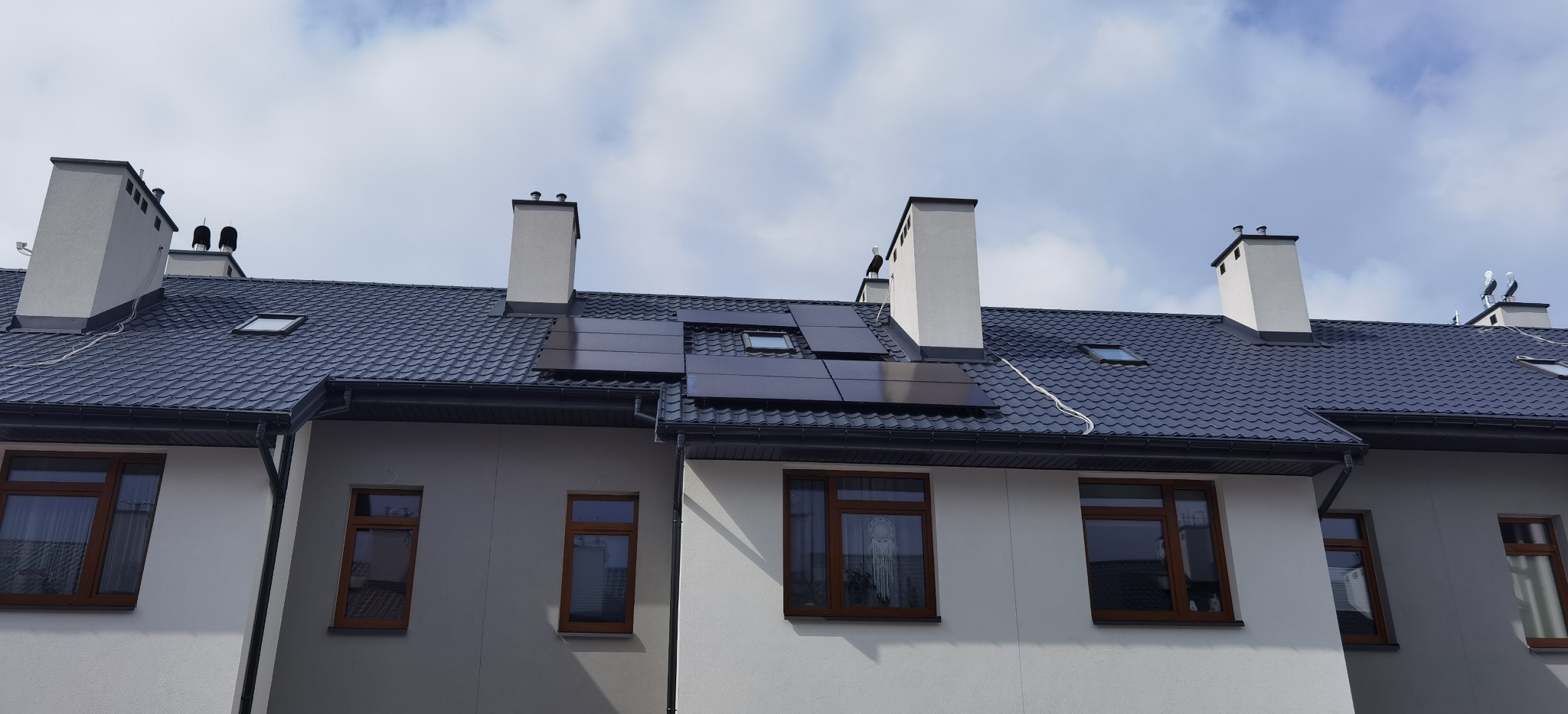 Rządowe domy szeregowe z ciemną dachówką i panelami słonecznymi, widok z dołu na błękitne niebo.