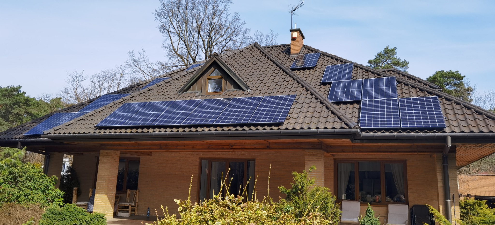 Dom jednorodzinny z brązową dachówką i panelami słonecznymi na dachu, z widocznym kominem i anteną telewizyjną, otoczony zielenią.