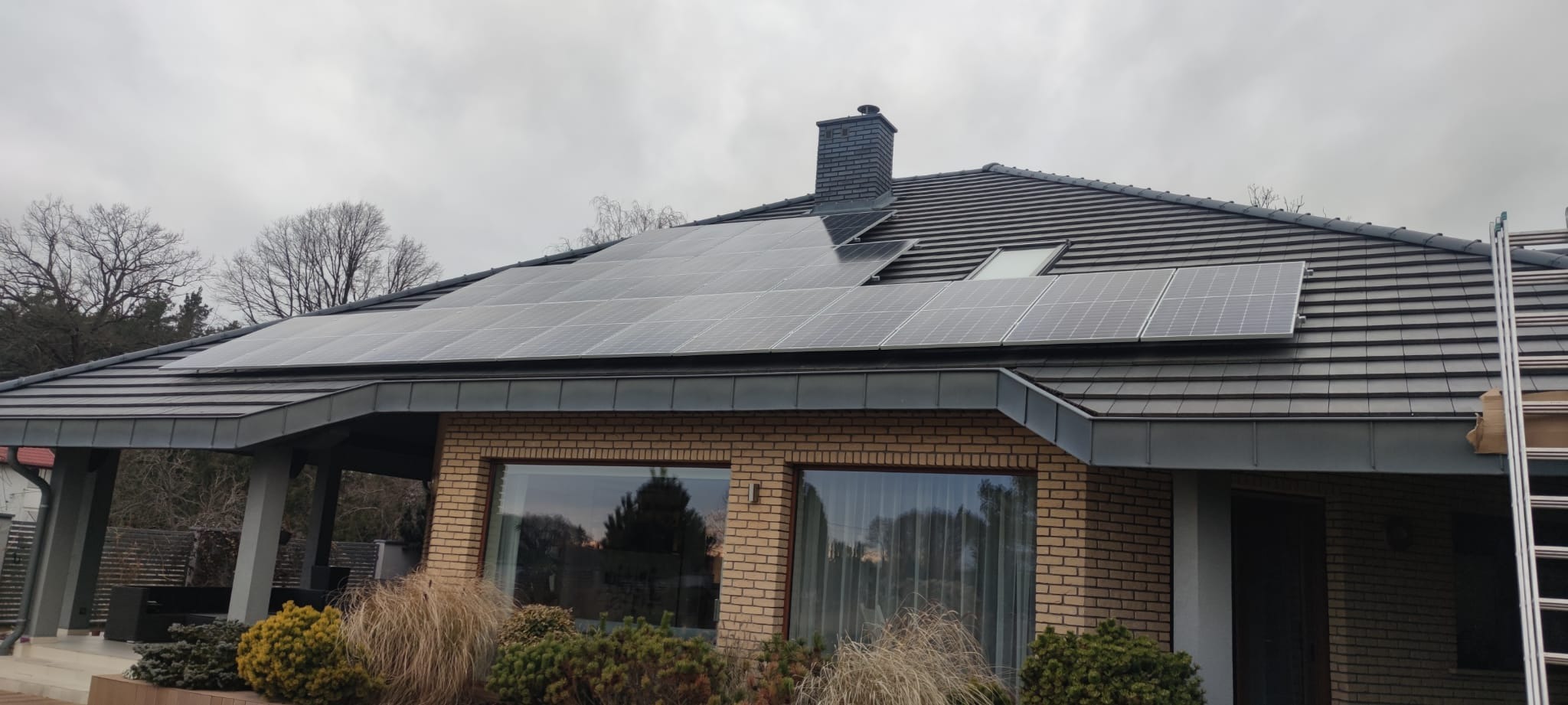 Panele słoneczne zamontowane na dachu domu z cegły, widoczne okna z zasłonami i fragment ogrodu z roślinami ozdobnymi, pochmurne niebo w tle.