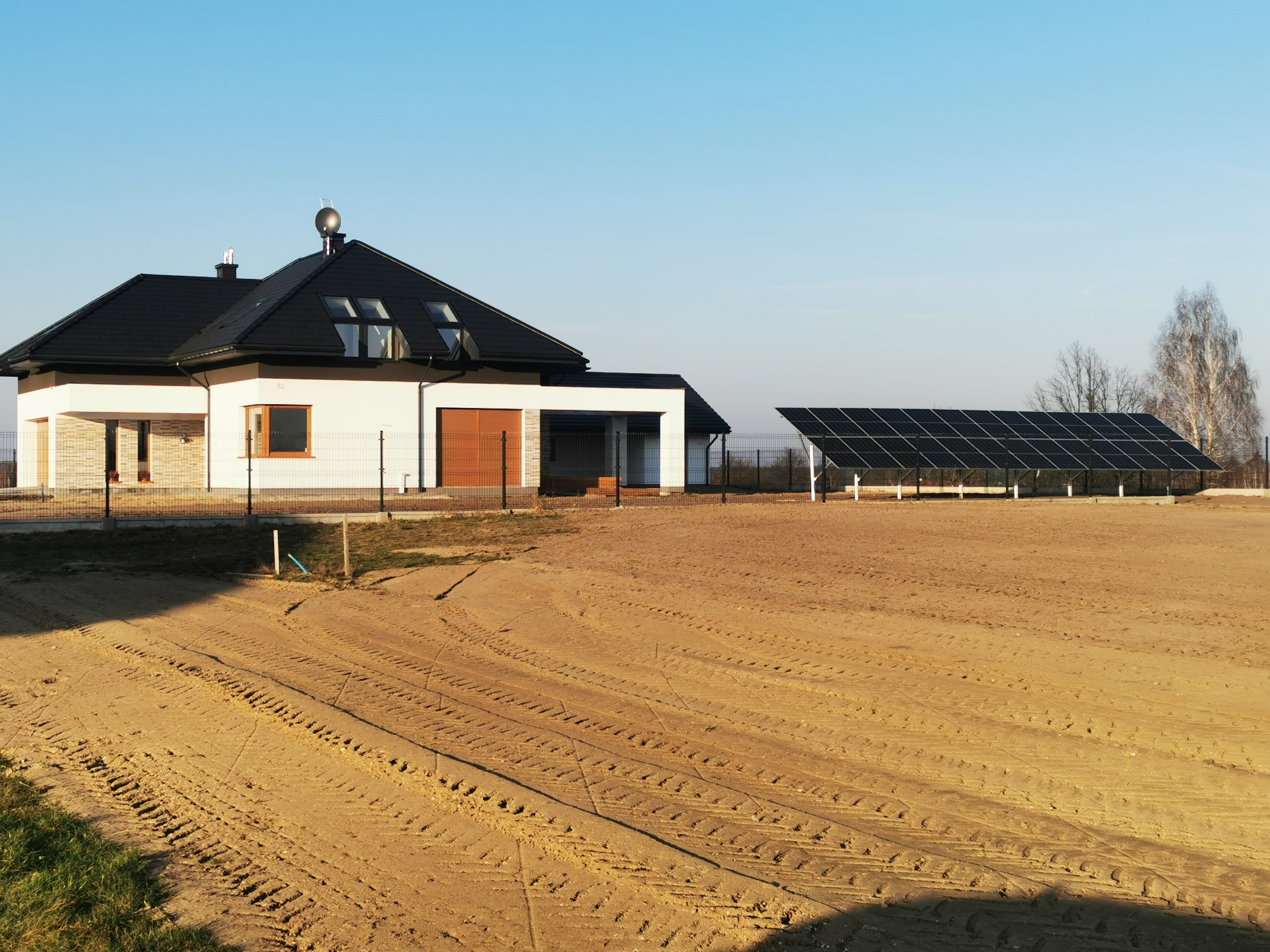 Nowoczesny dom jednorodzinny z czarnym dachem i panelami słonecznymi w tle, widok na rozległy teren z widocznymi śladami opon.