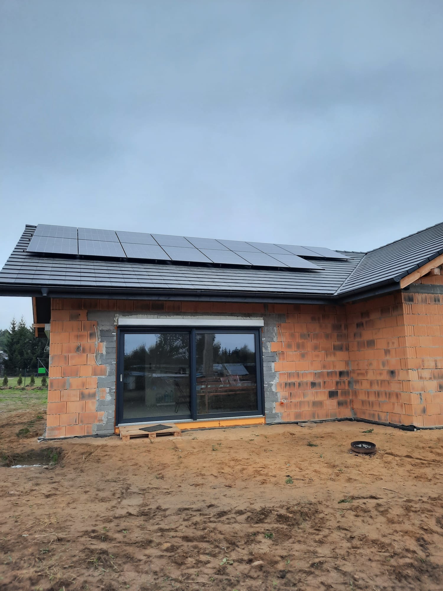 Niedokończony dom z czerwonej cegły z zamontowanymi panelami słonecznymi na dachu z dachówki ceramicznej, widoczne okno z szarą ramą i paleta pod nim, teren wokół domu piaszczysty.
