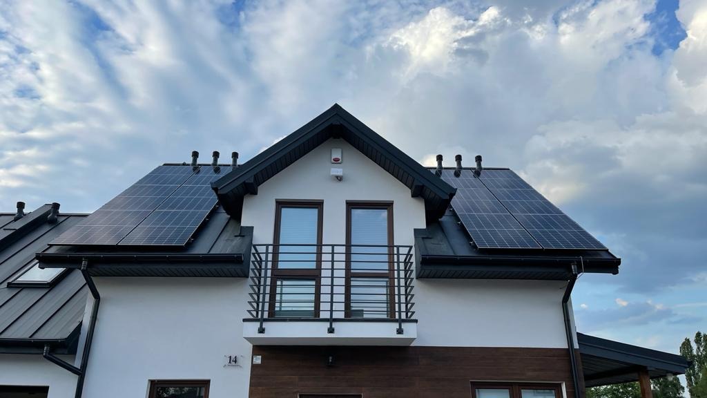 Nowoczesny dom jednorodzinny z zainstalowanymi panelami fotowoltaicznymi na dachu, widoczne okna balkonowe i elewacja w kolorze białym z elementami drewnopodobnymi.