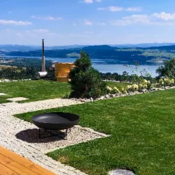 Ujęcie ogrodu z paleniskiem, brukowaną ścieżką i widokiem na jezioro, z drewnianą balią i kominem w tle.