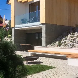 Nowoczesny dom z drewnianą elewacją i szarą kostką brukową na podwórku, z paleniskiem i niskimi ławkami.