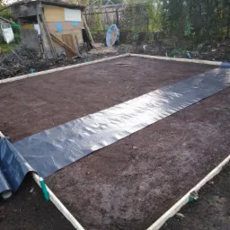 Przygotowanie terenu pod wylewkę fundamentów: szalunek z desek i rozłożona folia hydroizolacyjna na ubitej ziemi, widoczny barak i zarośla w tle.