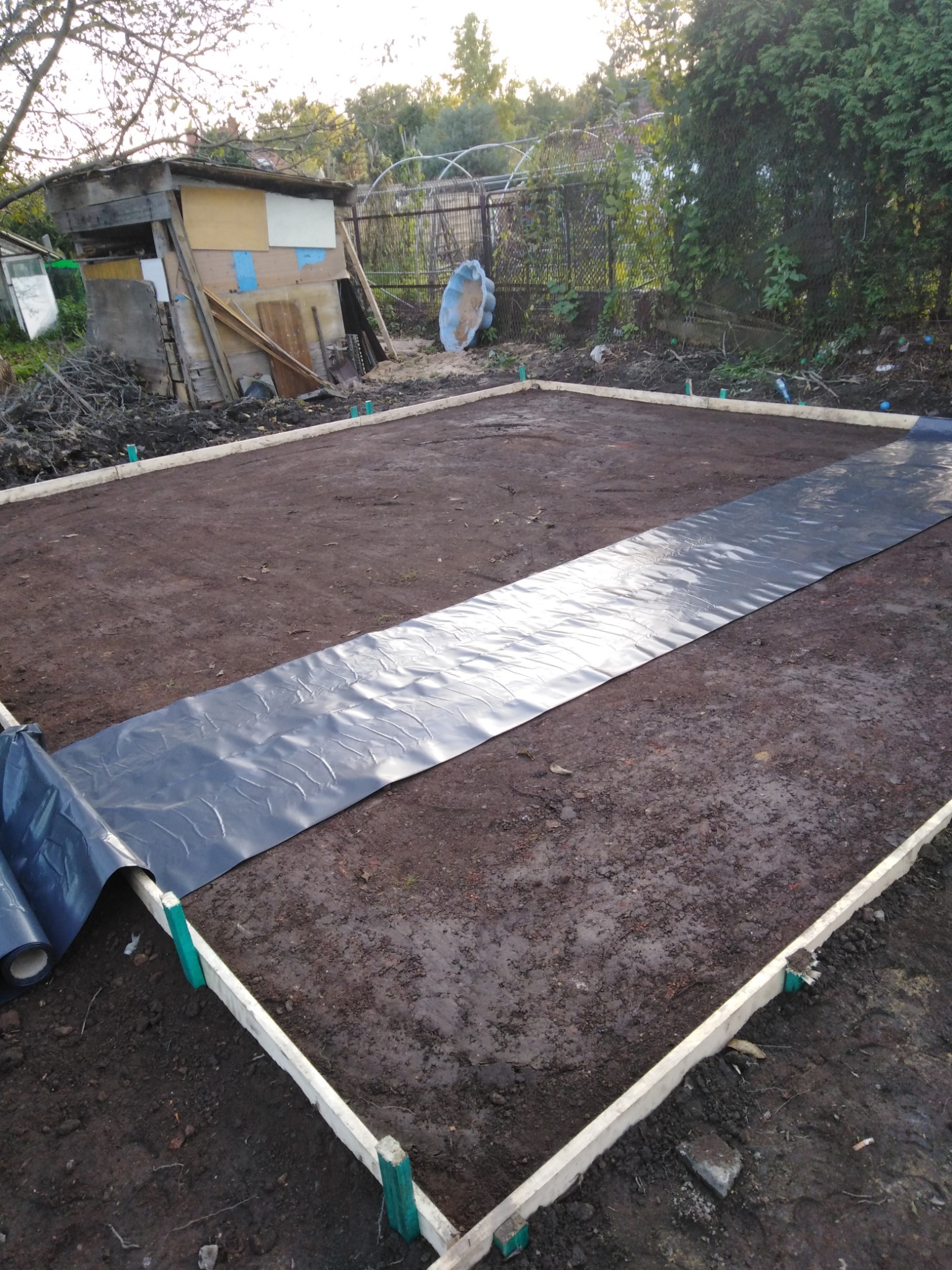Przygotowanie terenu pod wylewkę fundamentów: szalunek z desek i rozłożona folia hydroizolacyjna na ubitej ziemi, widoczny barak i zarośla w tle.