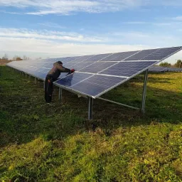 Instalator w ciemnej kurtce i czerwonych rękawicach, pochylony nad rzędem paneli słonecznych na łące, przy błękitnym niebie z obłokami.