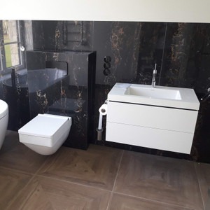 Nowoczesna łazienka z czarnymi marmurowymi płytkami, podwieszaną toaletą i białą szafką z umywalką. Widoczne chromowane elementy armatury i brązowe płytki podłogowe.