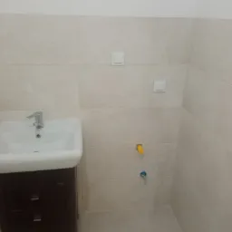 Wykończona łazienka z beżowymi płytkami, umywalką z szafką w kolorze ciemnego brązu, widoczne przyłącza wody z żółtą i niebieską zaślepką oraz dwa włączniki światła na ścianie.