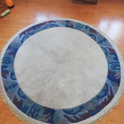 Wiktor Wasilczuk - Okrągły dywan z kremowym środkiem i niebiesko-fioletowym wzorem na obrzeżach, leżący na drewnianej podłodze, widoczny spryskiwacz i szczotka.