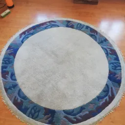 Okrągły dywan z kremowym środkiem i niebiesko-fioletowym wzorem na obrzeżach, leżący na drewnianej podłodze, widoczny spryskiwacz i szczotka.