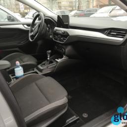 Wiktor Wasilczuk - Wnętrze samochodu z widocznymi fotelami z szarej tapicerki, czarną podłogą, deską rozdzielczą i butelką wody na siedzeniu pasażera. Logo firmy czyszczącej w prawym dolnym rogu.