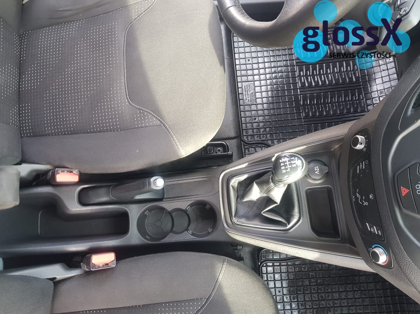 Wnętrze samochodu z widocznymi fotelami, konsolą środkową, dźwignią zmiany biegów i gumowymi dywanikami. Na dywaniku widoczne logo serwisu czystości GlossX.