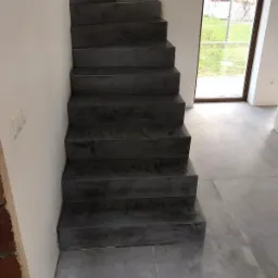 Betonowe schody wewnętrzne wykończone mikrocementem w surowym stylu, widok z dołu, szare płytki podłogowe i okno z widokiem na zewnątrz.