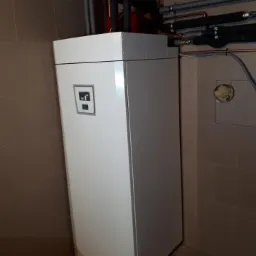 Biała pompa ciepła w rogu pomieszczenia z brązowymi płytkami, widoczne rury i manometr