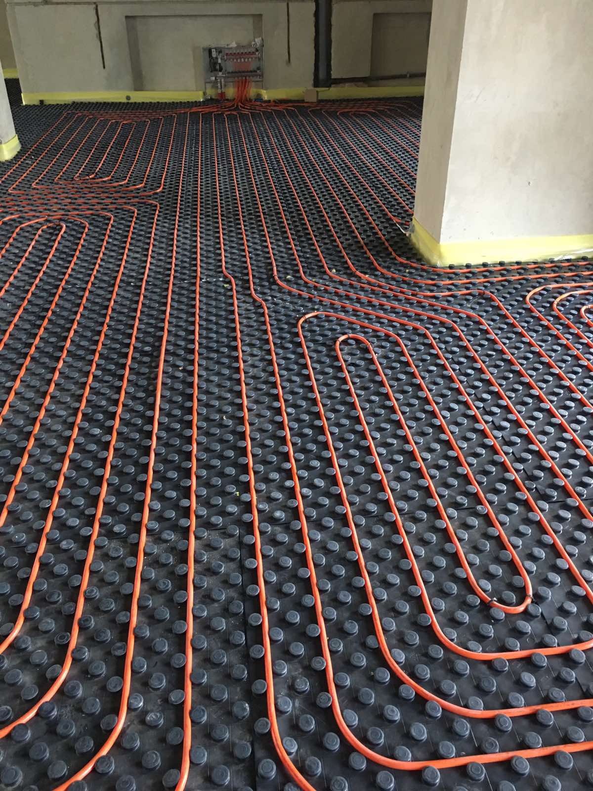 Pomarańczowe rury ogrzewania podłogowego ułożone na czarnej izolacji z wypustkami, widok z góry na dużą powierzchnię w surowym wnętrzu budynku w trakcie budowy.