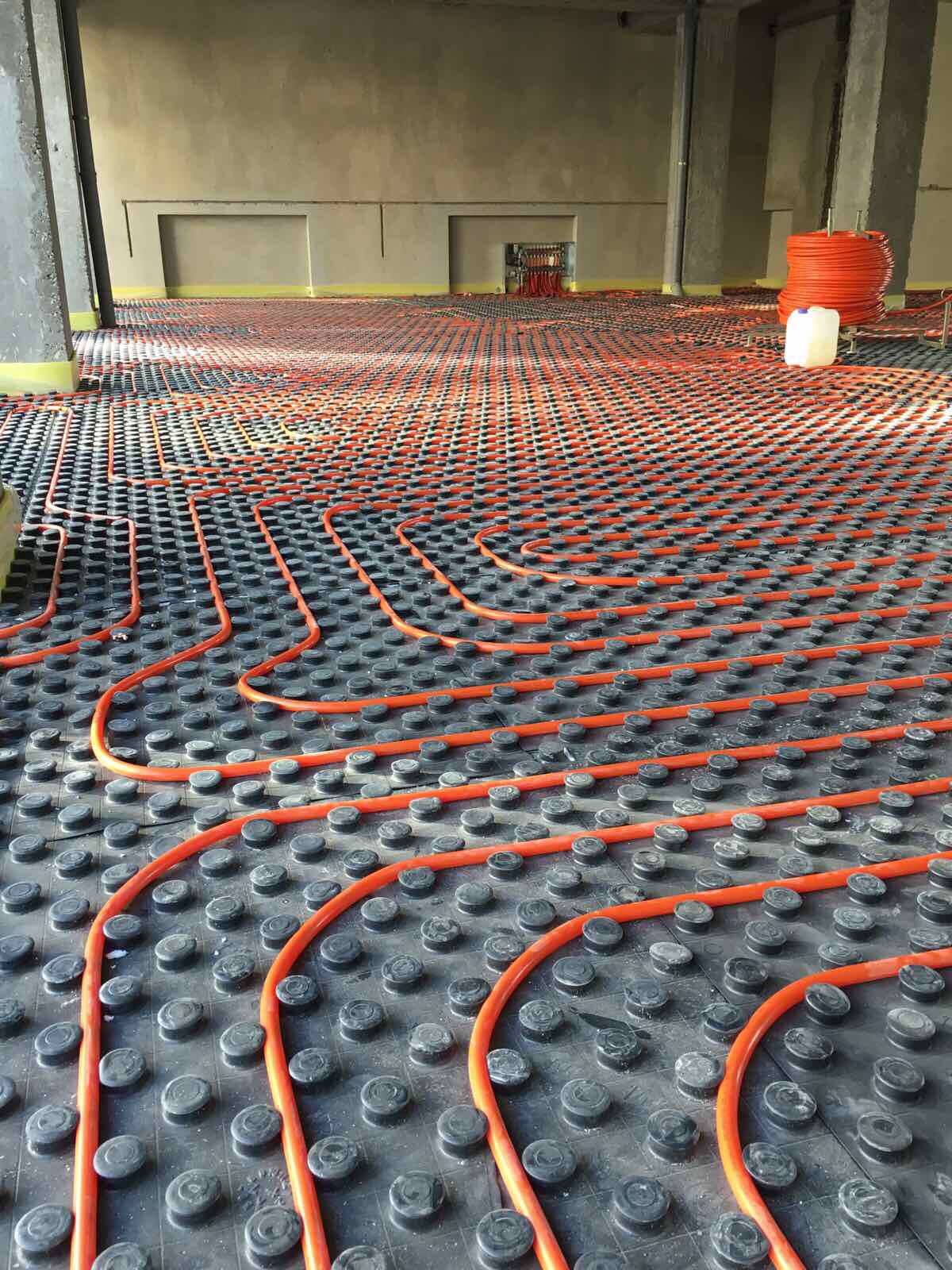 Pomarańczowe rury ogrzewania podłogowego ułożone na czarnej izolacji z wypustkami w surowym wnętrzu budynku, widoczna rolka rur i biały zbiornik w tle.