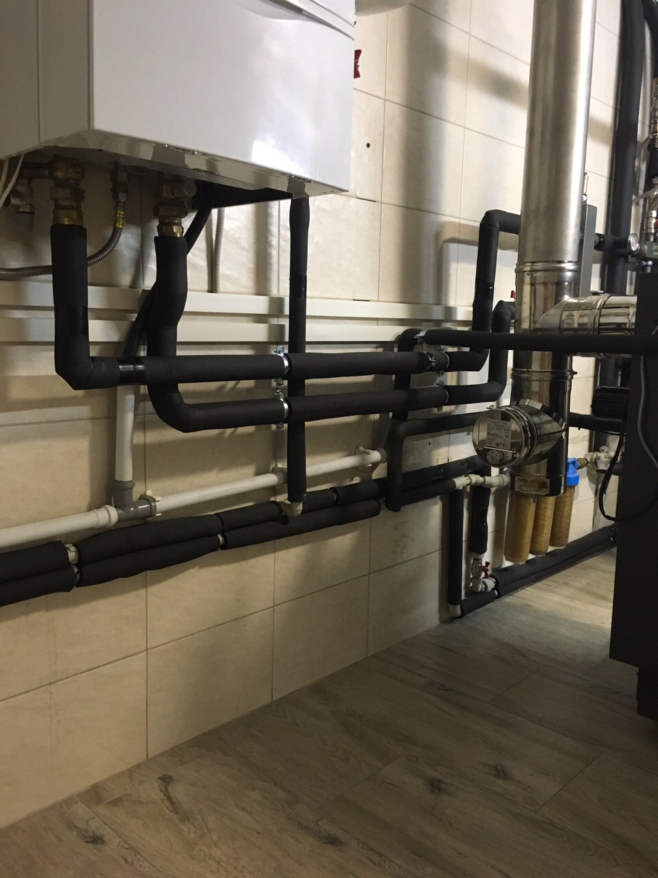 Biały piec grzewczy z czarnymi rurami izolowanymi termicznie, zamontowany na kafelkowanej ścianie w kotłowni z drewnopodobną podłogą.