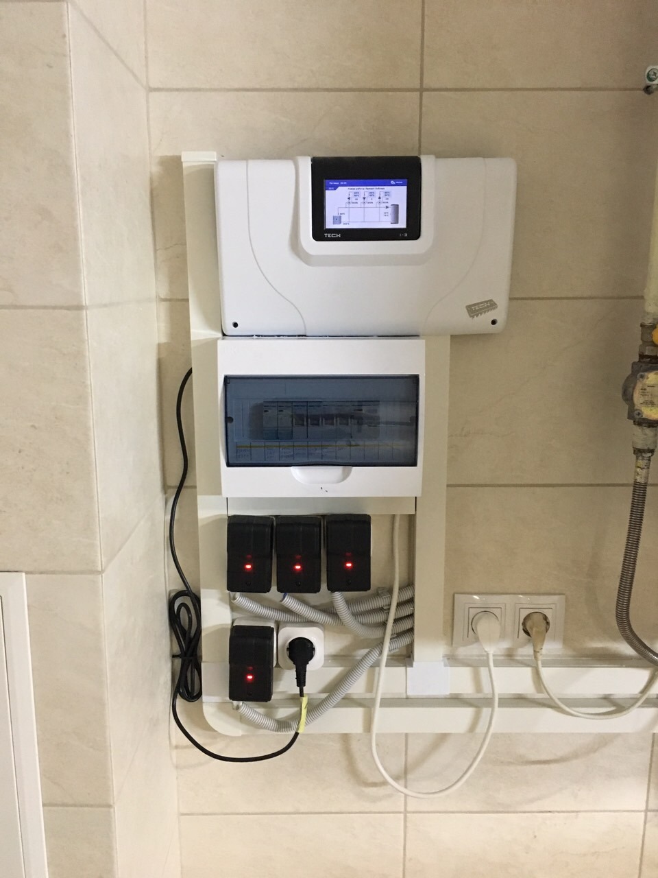 Panel sterowania z wyświetlaczem, rozdzielnica elektryczna i czujniki temperatury zamontowane na ścianie z beżowych płytek. Widoczne przewody i gniazdka elektryczne.