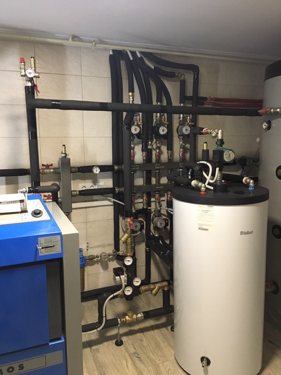 Złożona instalacja grzewcza z kotłem na paliwo stałe, zbiornikiem akumulacyjnym Vaillant i licznymi rurami z izolacją, manometrami i zaworami w kotłowni z beżowymi płytkami ściennymi i podłogą...