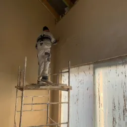 Pracownik na rusztowaniu aplikuje gładź gipsową na ścianę w surowym wnętrzu, widoczne drewniane elementy konstrukcji dachu i folia ochronna na oknie.