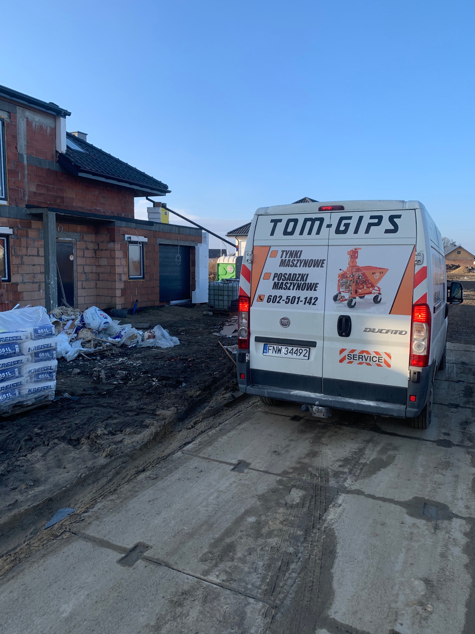 Furgonetka firmy Tom-Gips z reklamą tynków i posadzek maszynowych na tle budowy w stanie surowym otwartym i stert worków z zaprawą.