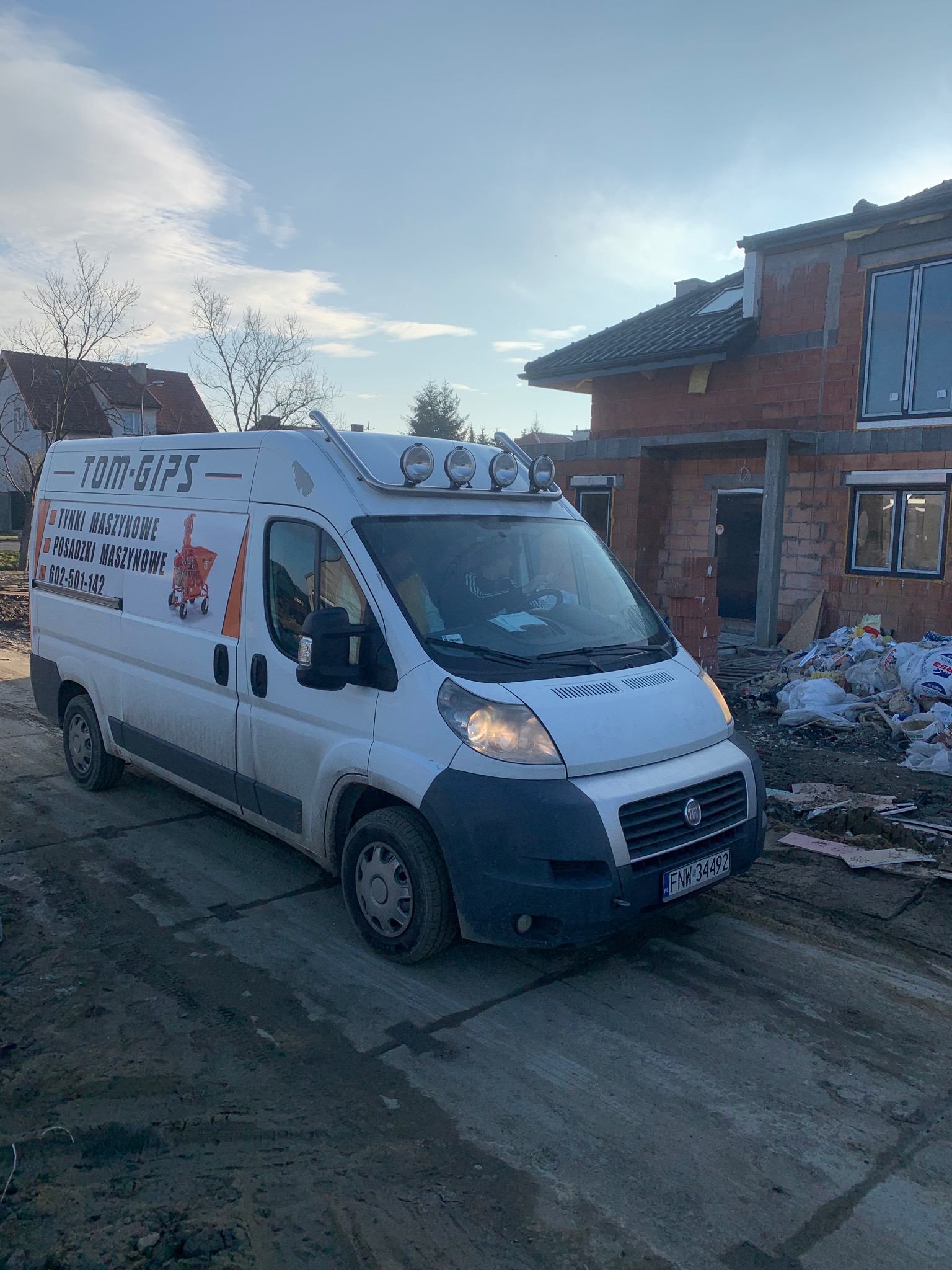 Brudny biały bus firmy Tom-Gips z reklamą tynków i posadzek maszynowych, zaparkowany na placu budowy domu w stanie surowym otwartym.