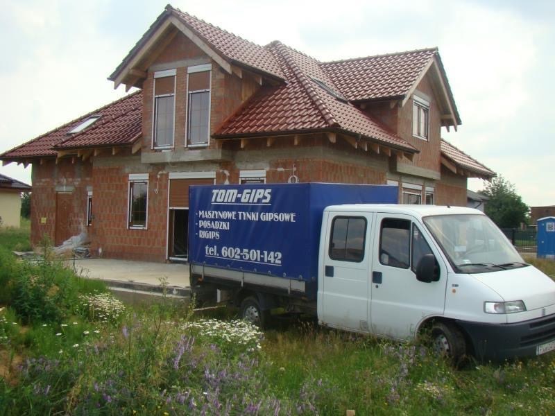 Nowo budowany dom z czerwonej cegły, z częściowo zamontowaną stolarką okienną, w tle biały samochód dostawczy z reklamą firmy wykonującej maszynowe prace gipsowe.