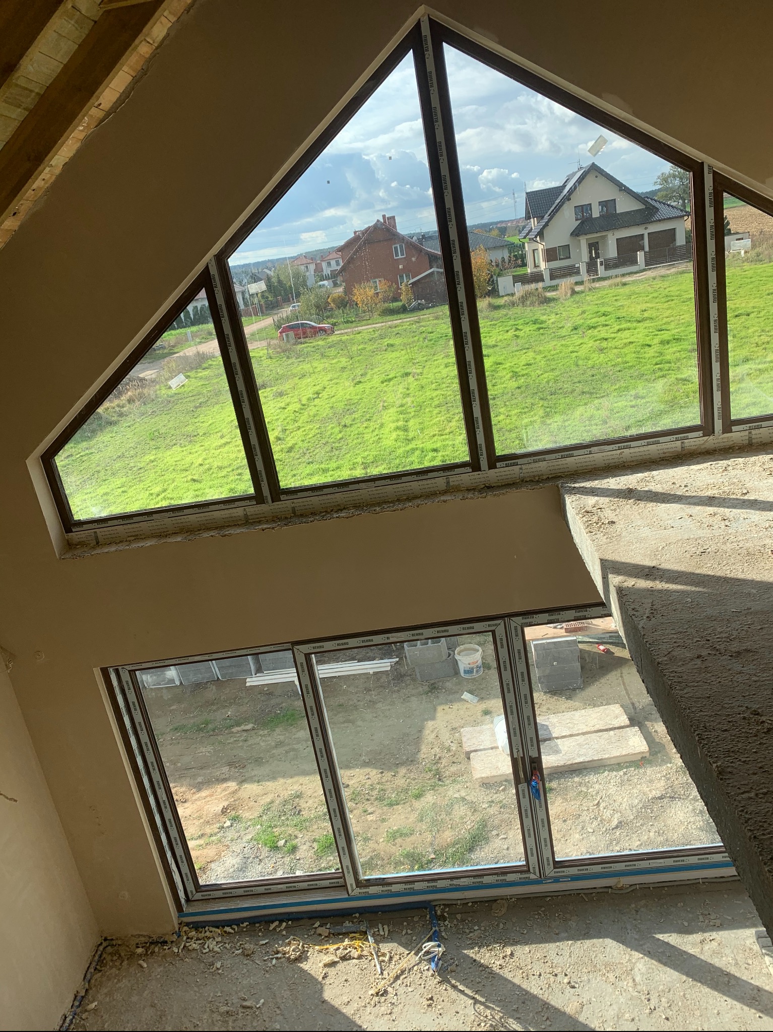 Wnętrze w trakcie prac wykończeniowych: świeżo otynkowana ściana pod skośnym oknem z widokiem na domy i zielony trawnik, widoczne materiały budowlane i fragment schodów.