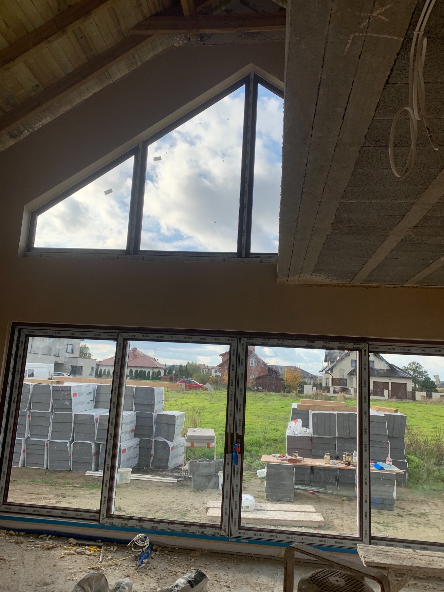 Wnętrze w surowym stanie z dużym oknem trójkątnym pod sufitem i panoramicznym oknem z widokiem na posesję z ułożonymi bloczkami betowymi i narzędziami.