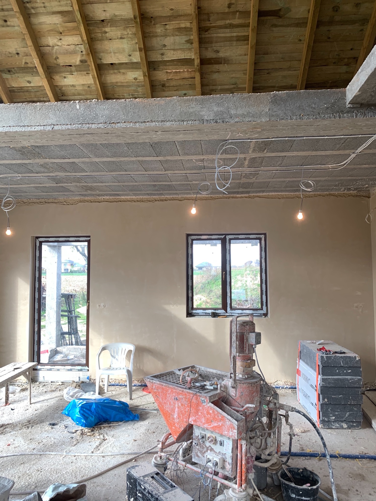 Wnętrze budynku w trakcie prac wykończeniowych, ściany pokryte świeżo nałożonym tynkiem, widoczna maszyna do natrysku tynku oraz stos płyt izolacyjnych, surowe oświetlenie z żarówek na kablach.