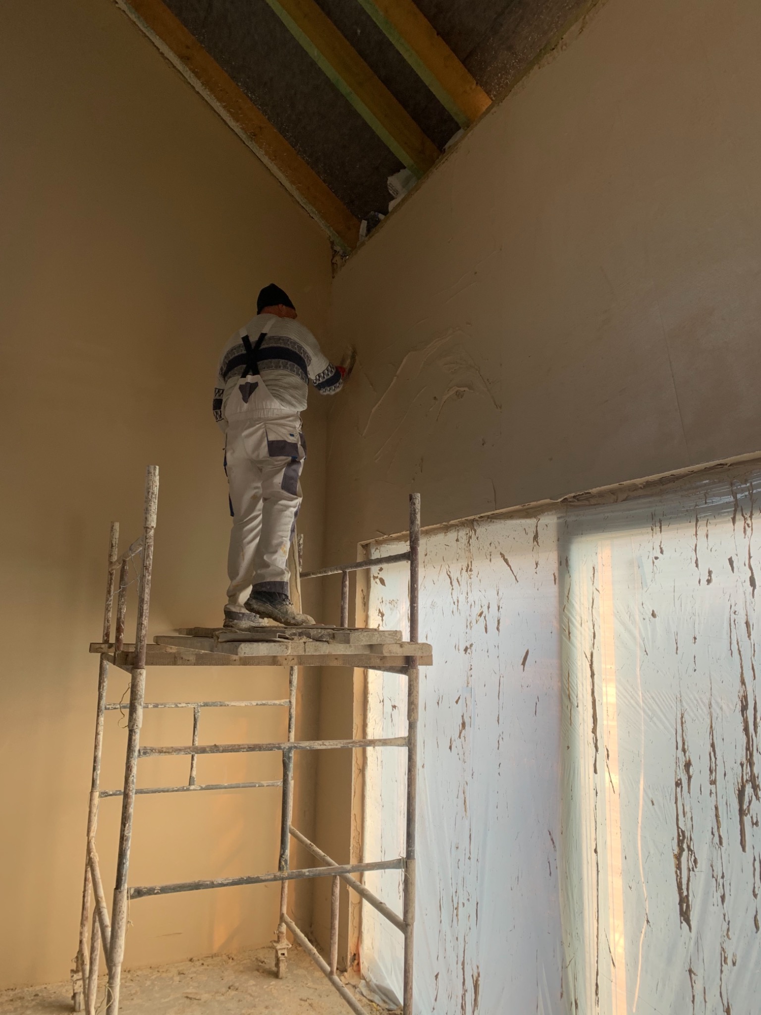 Pracownik na rusztowaniu aplikuje gładź gipsową na ścianę w surowym wnętrzu, widoczne drewniane elementy konstrukcji dachu i folia ochronna na oknie.