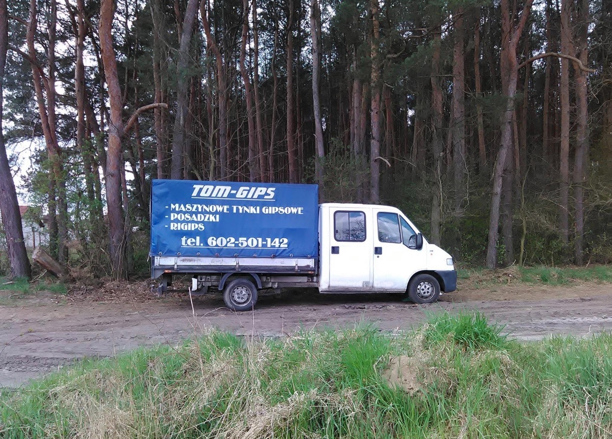 Biała furgonetka z niebieską plandeką z napisem 'TOM-GIPS' i ofertą usług tynkarskich, zaparkowana na polnej drodze na skraju lasu.