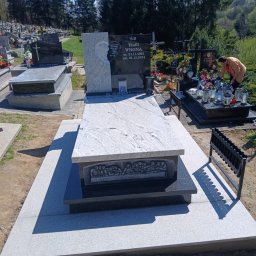 K2 STONE Kamieniarstwo Chruślicki - Granitowy nagrobek z płaskorzeźbą Ostatniej Wieczerzy na cmentarzu, w tle drzewa i inne groby. Osoba w oddali. Jasny dzień, błękitne niebo.